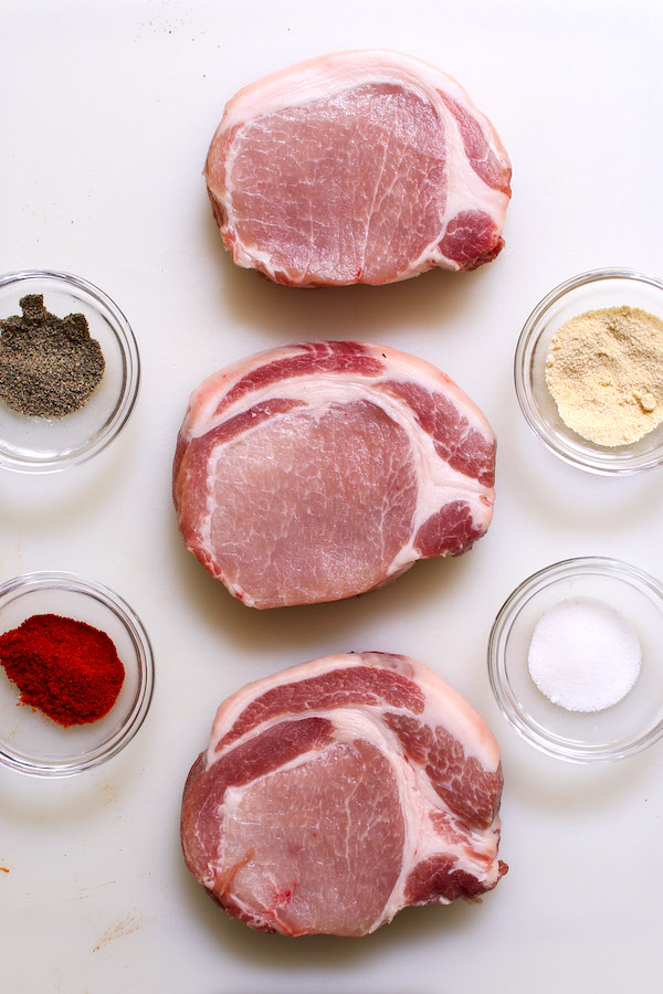 Raw boneless pork chops alongside 4 simple seasonings: paprika, garlic powder, salt and pepper