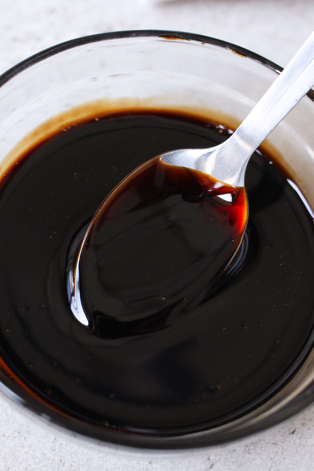 Balsamic reduction in a small bowl with a spoon showing the thick, glaze-like texture that is perfect for drizzling and coating meats, seafood, vegetables, salads, fruits and more