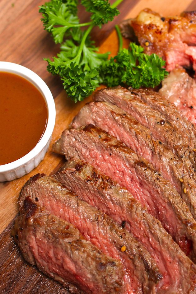 Beautiful broiled ribeye steak sliced thinly on a carving board with steak sauce and a sprig of parsley on the side