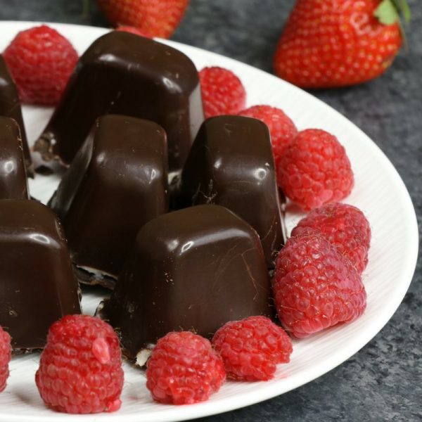 This photo shows chocolate covered cheescake bites arranged on a serving plate surrounded by fresh raspberries