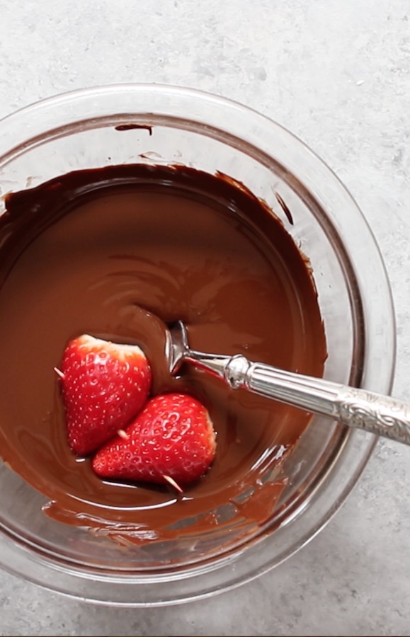 This photo shows dipping heart shaped strawberries into melted semisweet chocolate in a bowl to make Chocolate Covered Strawberries.