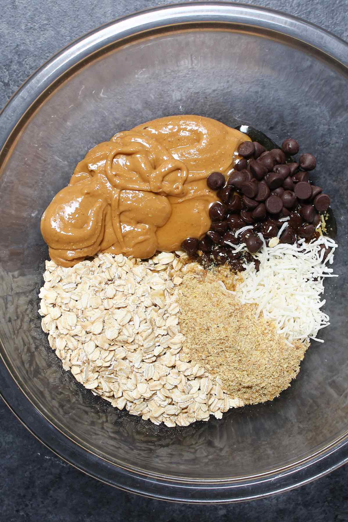 Ingredients used in an energy balls recipe in a mixing bowl: rolled oats, peanut butter, flaxseed meal, shredded coconut, chocolate chips, maple syrup and vanilla