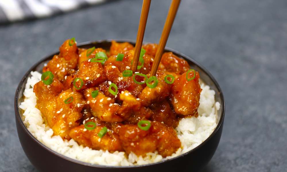 General Tso Chicken Crock Pot - this photo shows this dish served on top of white rice and garnished with minced green onion and sesame seeds, with chopsticks going in to pick up a piece. A tangy and flavorful slow cooker recipe to feed the entire family.