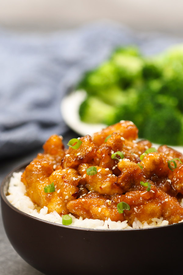 General Tso's Chicken - this photo shows a closeup of crispy chicken pieces in a sticky sauce on top of white rice served in a bowl with fresh broccoli in the background