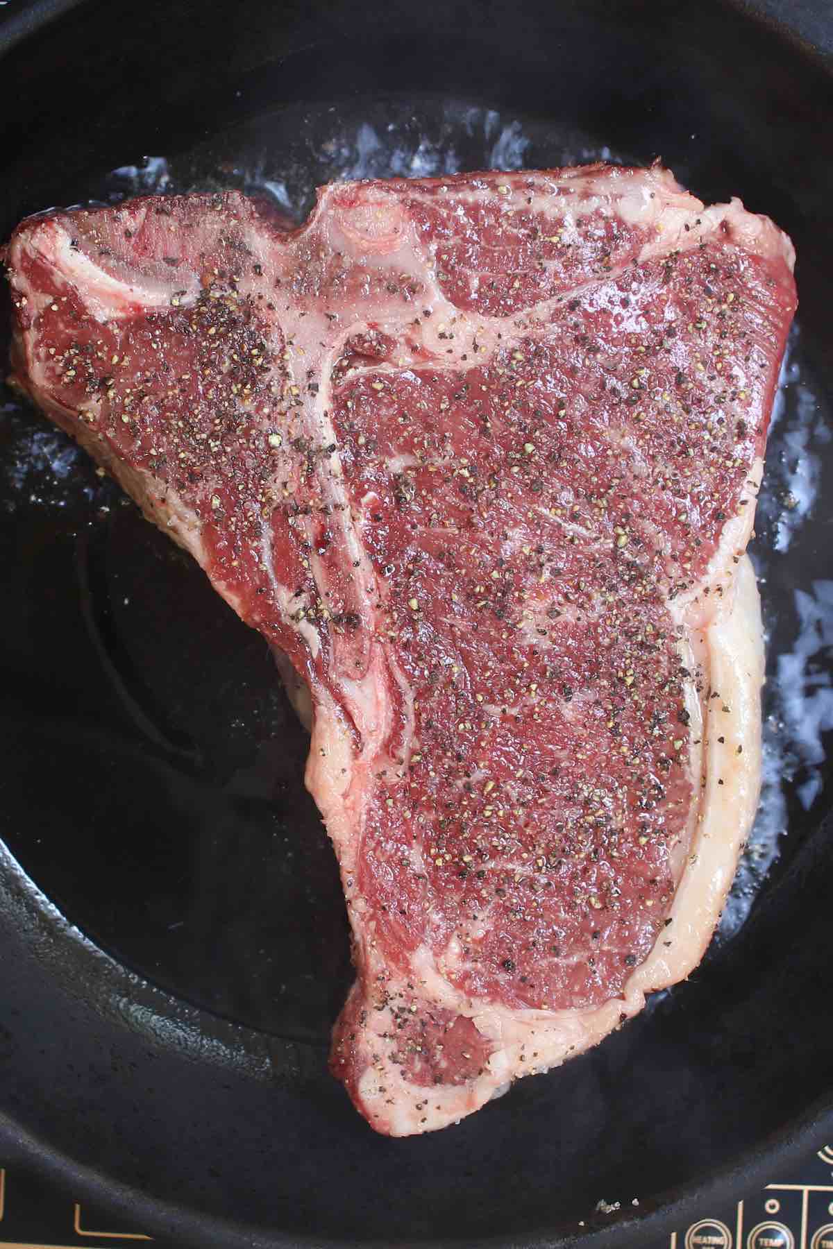 Overhead view of cooking a seasoned t-bone steak in a hot cast iron skillet with sizzling all around