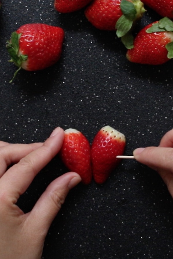 This photo shows fastening two strawberry halves together sideways with a toothpick to make heart-shaped Chocolate Covered Strawberries Then place the two halves next to each other and form a heart shape.