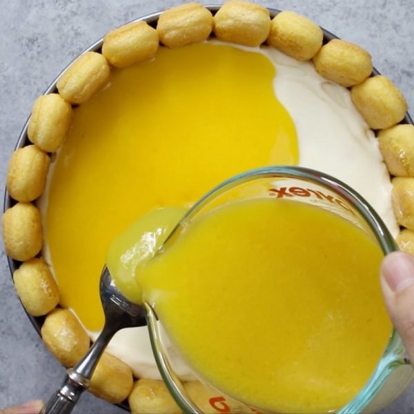 This photo shows adding a mango glaze to the top of a mango cheesecake charlotte by pouring from a measuring cup onto the back of a spoon to create a dribble