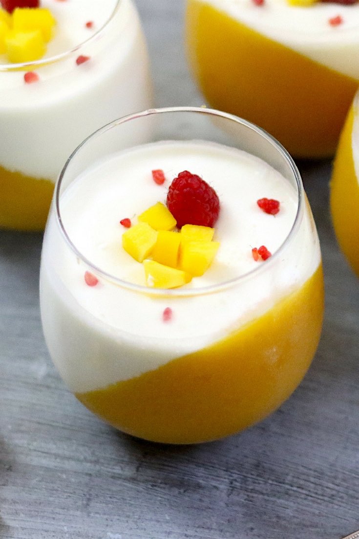 Mango panna cotta served in stemless wine glasses and garnished with raspberries and chunks of fresh mango