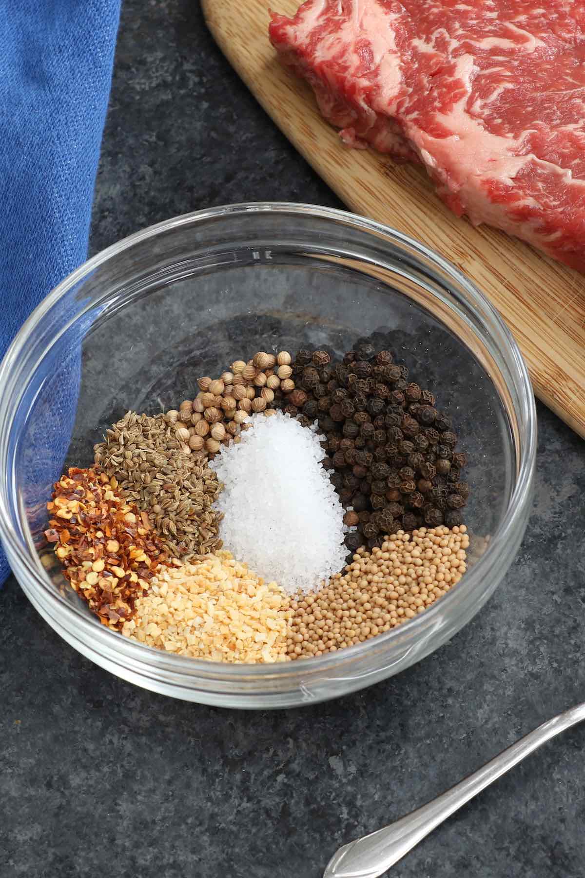 Whole spices in a bowl including black peppercorns, coarse salt, coriander seeds, anise seeds, mustard seeds, granulated garlic and onion and red pepper flakes