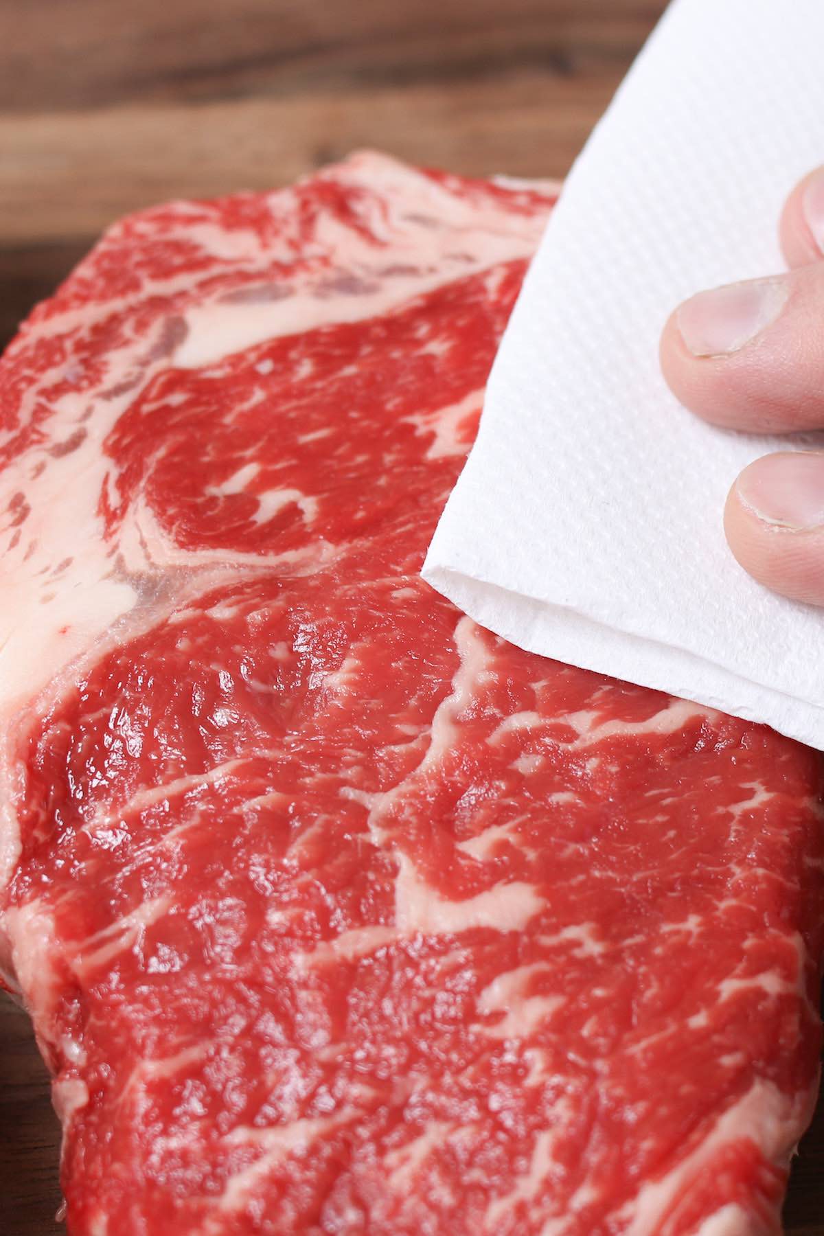 Closeup of pat drying a rib eye steak with paper towels to remove excess moisture that could interfere with searing