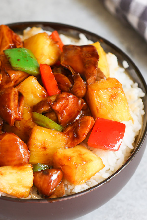 Pineapple Chicken featuring chunks of chicken thighs, fresh pineapple, red bell pepper and green bell peppers on a red of rice in a serving bowl