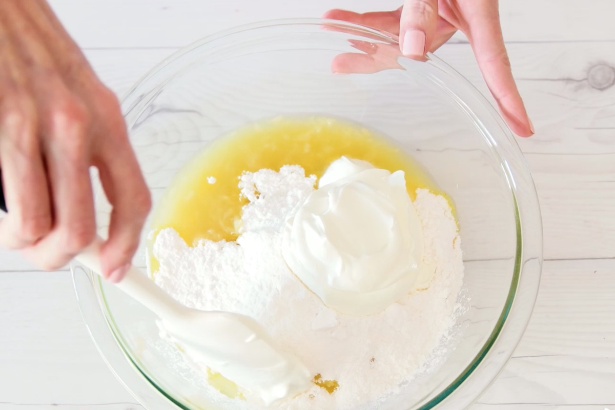 Mixing crushed pineapple, vanilla pudding mix, and sour cream in a clear bowl on the counter. 