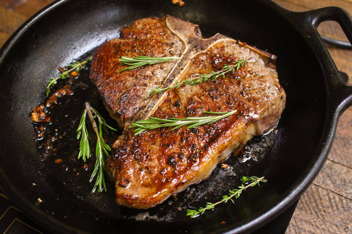 Porterhouse Steak in a cast iron pan after being seared and oven baked to caramelized perfection with salt, pepper, garlic, butter and sprigs of fresh rosemary and thyme 