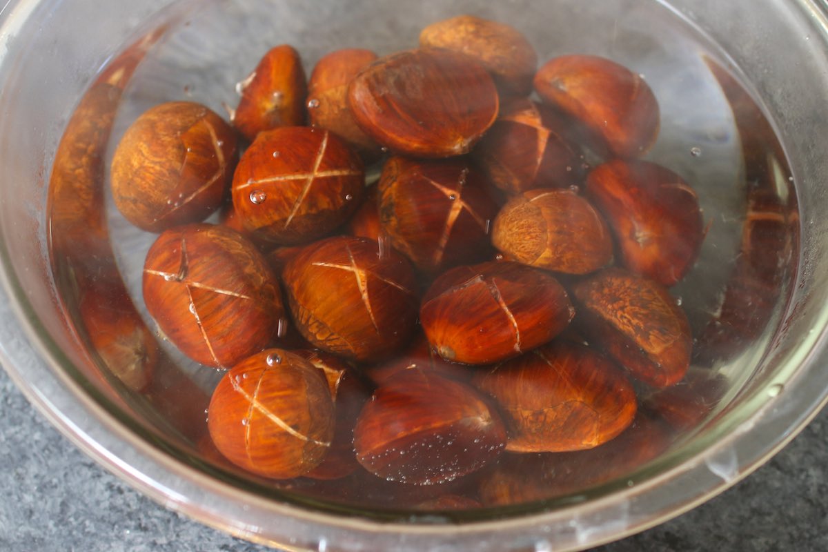 Soaking scored raw chestnuts in hot water in a clear bowl.