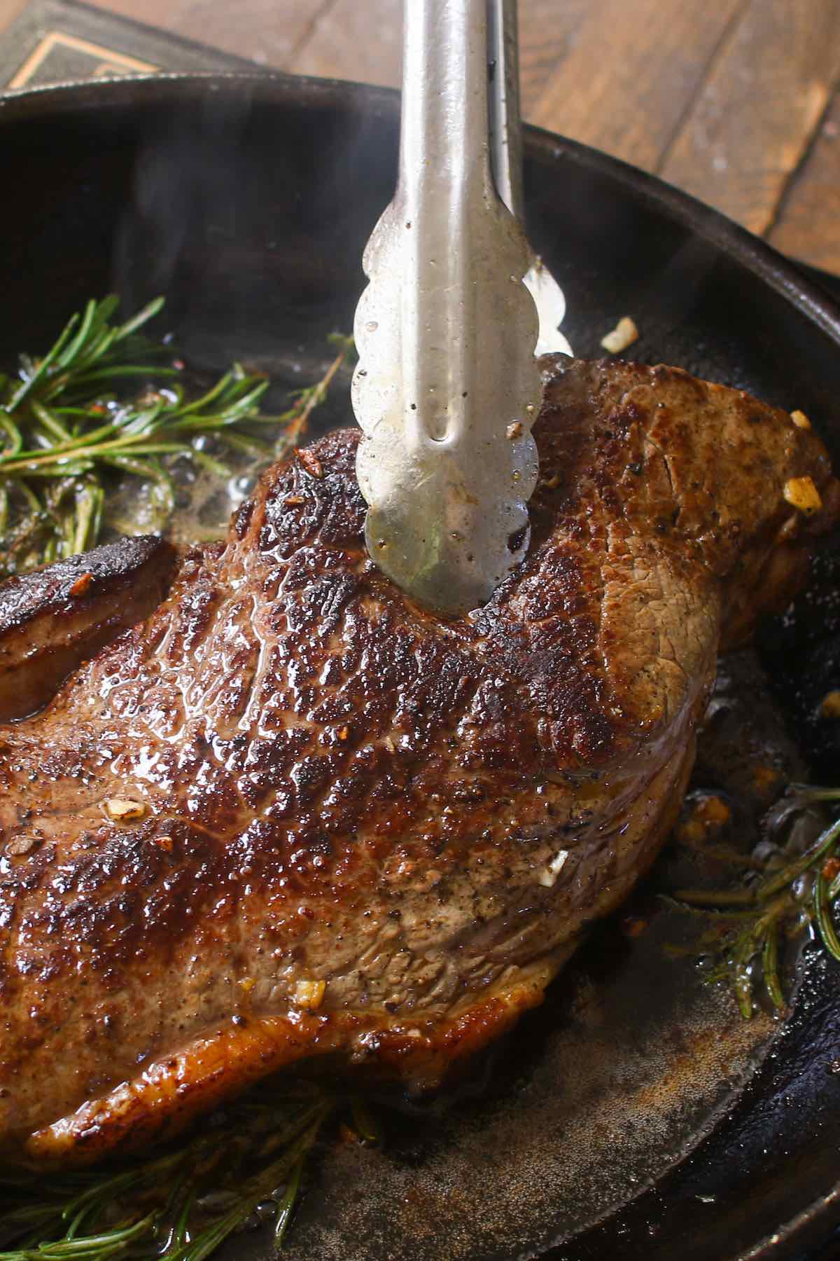 Flipping a steak in a cast iron pan using kitchen tongs