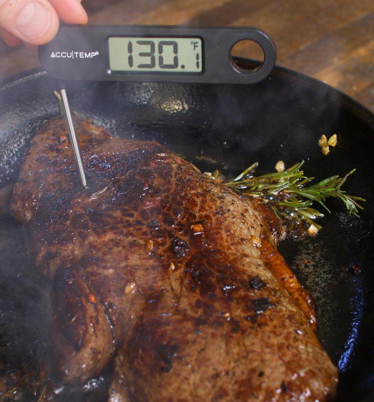 Checking doneness of a steak in a cast iron pan by inserting an instant-read thermometer into the thickest section of the meat. The reading of 130°F indicates medium doneness.