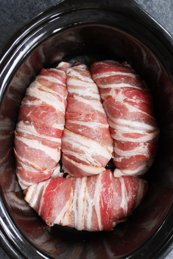 Slow cooking chicken breasts that have been rubbed with seasonings and wrapped in bacon before being placed in a single layer in the slow cooker for cooking