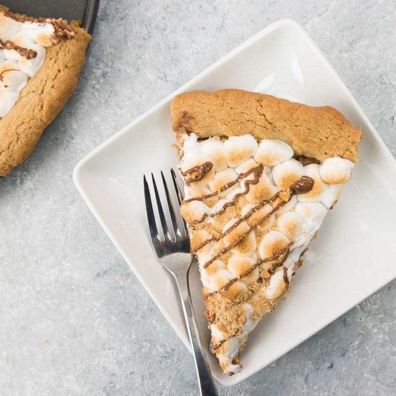 This photo shows a piece of Smores Pizza served on a white dessert plate with a fork next to the pizza pan