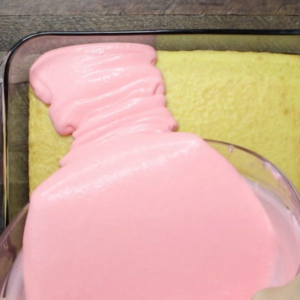 This is a photo showing how to pour strawberry jello filling onto the yellow cake layer in the baking dish as part of the steps to make strawberry jello cake