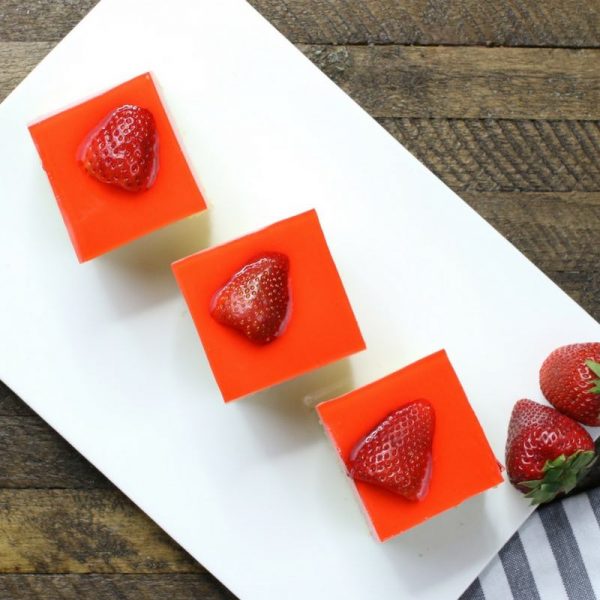 Three square pieces of strawberry jello cake served on a rectangular white plate and garnished with fresh strawberries on the side