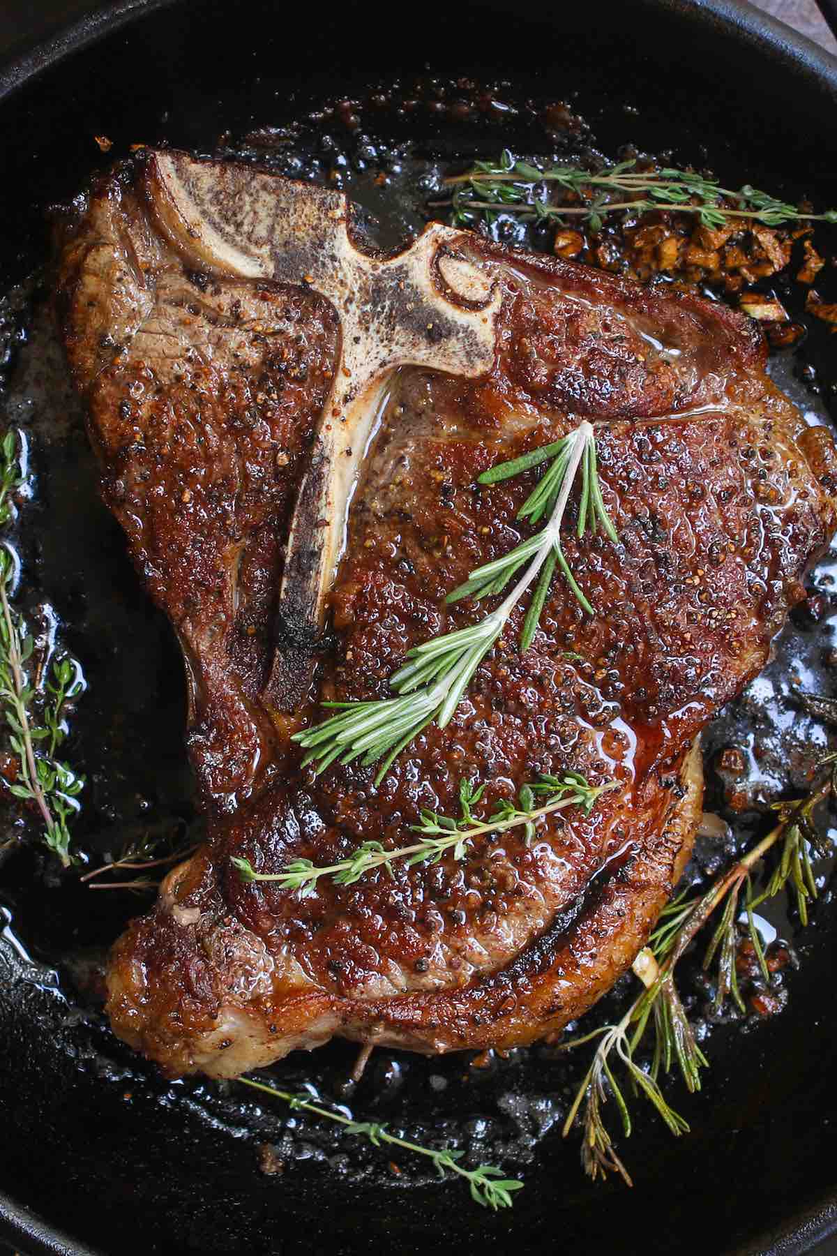 Pan seared T-bone Steak in a cast-iron pan showing a beautiful golden crust with sprigs of fresh rosemary and thyme on top for garnish