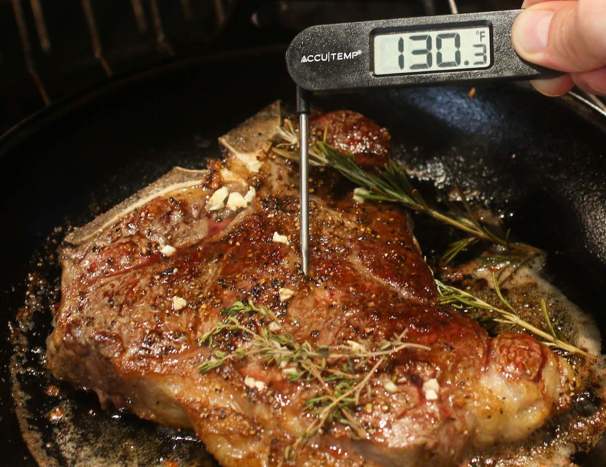 Checking doneness of a t-bone steak by inserting an instant-read thermometer into the middle of the striploin section about an inch from the bone. The reading of 130 degrees F indicates the steak is cooked medium rare