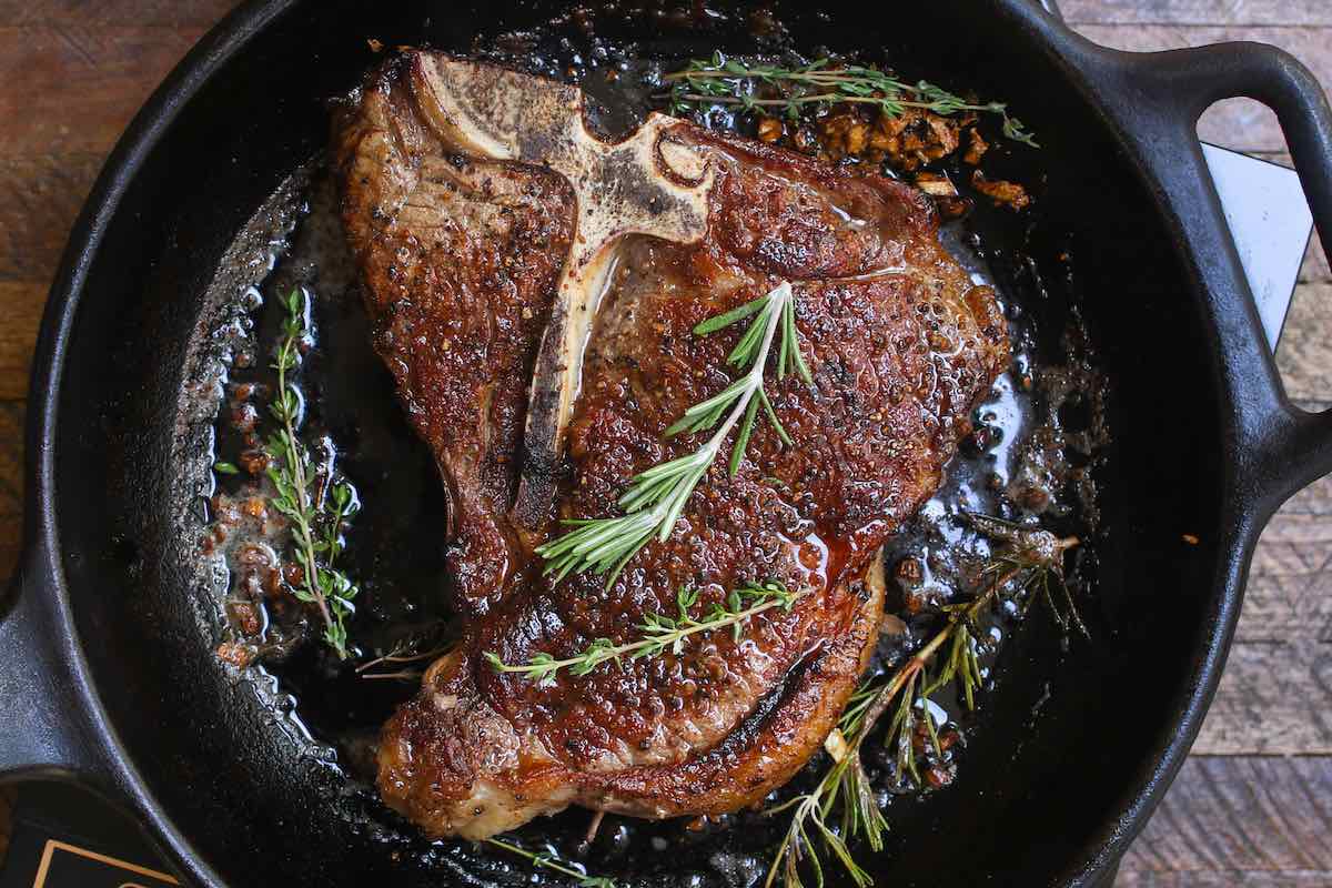 Overhead view of searing a t-bone steak in a very hot cast iron frying pan with the fat sizzling around the meat